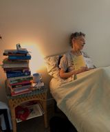 a woman laying on a bed surrounded by piles of books, a book opened against her chest as if she had fallen asleep while reading it